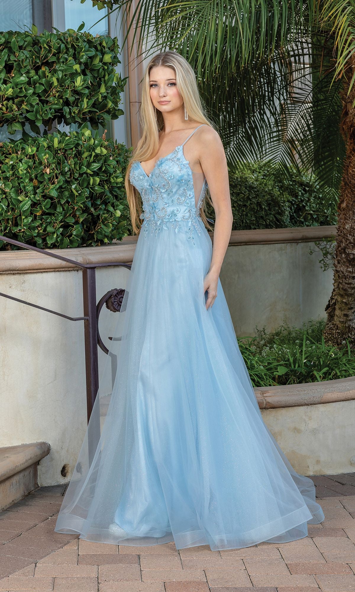 A woman in the Long A-Line Tulle Prom Ball Gown with Flowers 4276 stands outdoors on a stone walkway, surrounded by greenery and palm trees. The light blue dress features floor-length tulle and 3-D floral designs.